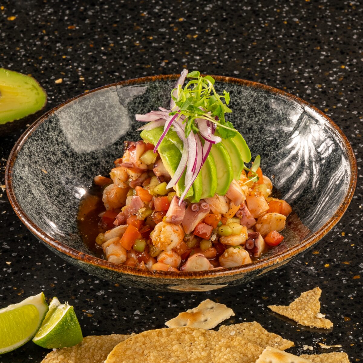 Shrimp and octopus ceviche in a stone bowl, topped with sliced avocado, red onion and microgreens, with tostadas and lime