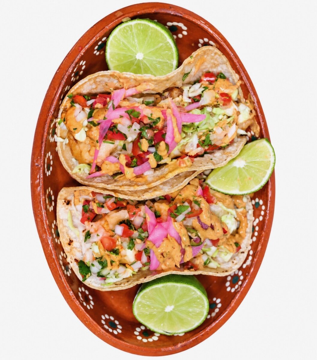 Two fish tacos on a traditional clay plate, topped with pico de gallo, pickled red onion, cilantro and chipotle sauce, with lime wedges
