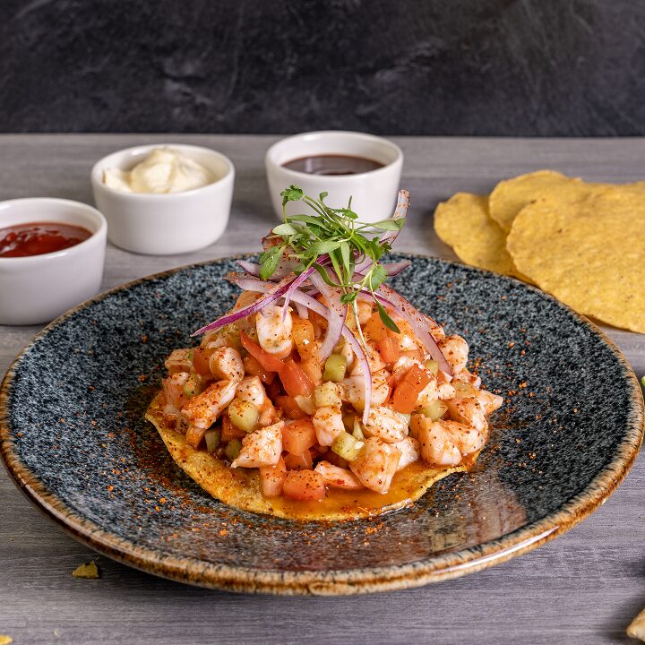 Shrimp tostada topped with red onion and microgreens on a speckled plate, with salsas and tostadas behind